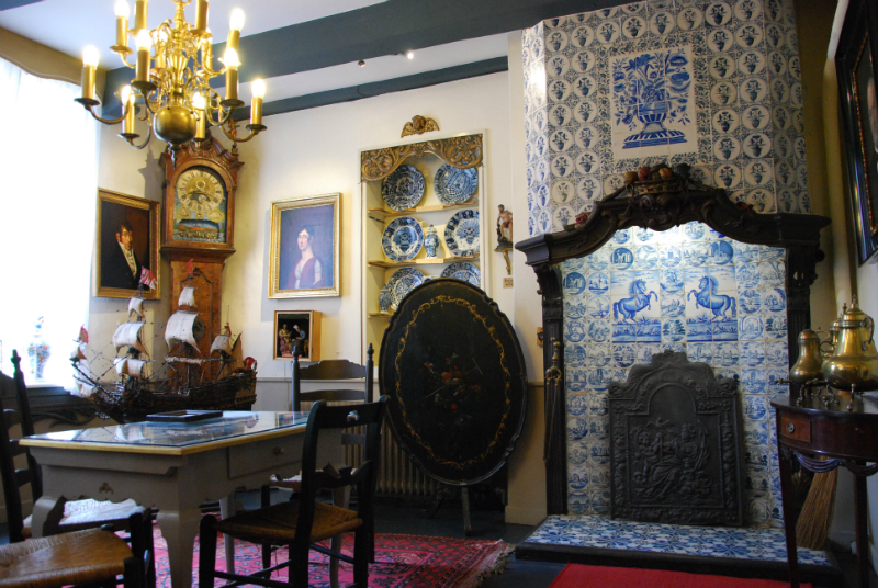 Museum: Geschm&uuml;ckter Raum mit gefliestem Kamin, Schmucktellern im Regal, Kronleuchter, Standuhr