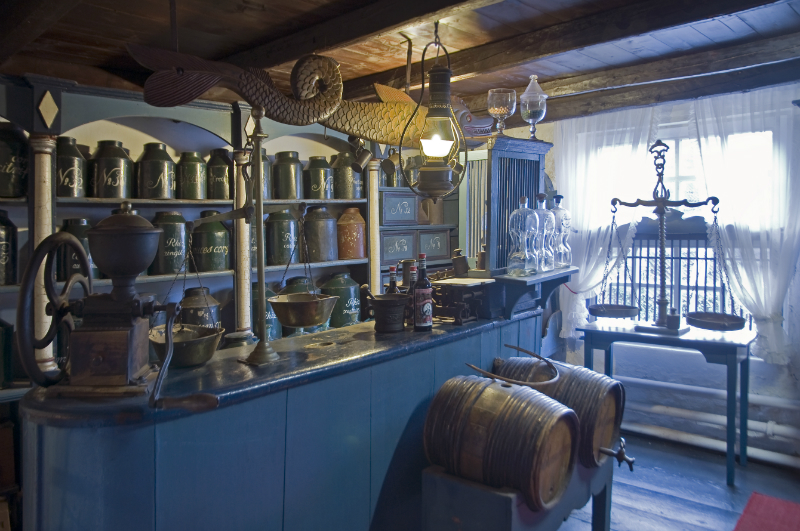 Museum: Historische Ladenansicht. Eine Verkaufstheke mit antiken Waagen, Holzf&auml;ssern, Teedosen und traditionellen Apothekengl&auml;sern, eingerahmt von rustikalen Holzbalken.